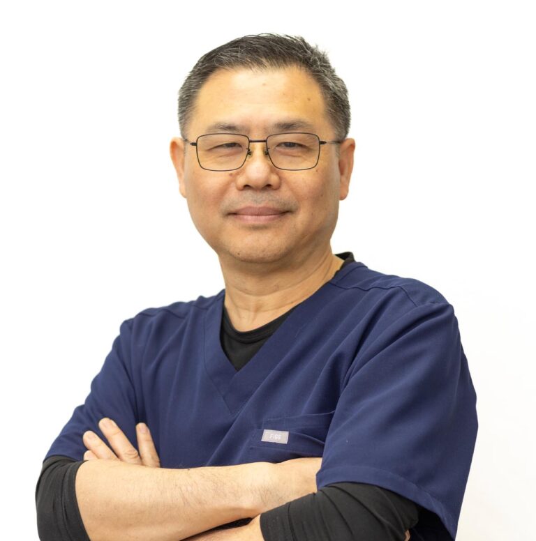 Headshot of Doctor Thomas Moe wearing a dark blue scrub top with a black undershirt, their arms folded across their chest. The outfit includes a small tag on the left, against a plain white backdrop.