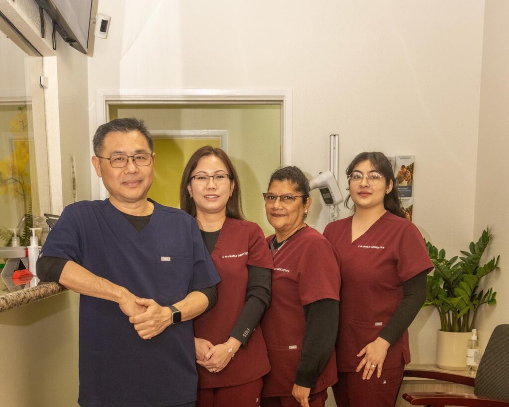 Doctor Thomas Moe with his team at c w family dentistry in a medical or dental office. Three are dressed in maroon scrubs and one in navy blue, all standing near a doorway. The setting includes a counter with items, a wall-mounted plant, and medical equipment, emphasizing a professional healthcare environment.